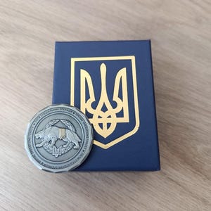 Może przedstawiać: Srebrna moneta z wizerunkiem lwa i tekstem cyrylicą, obok granatowego pudełka ze złotym emblematem. Całość umieszczona na jasnej drewnianej powierzchni.