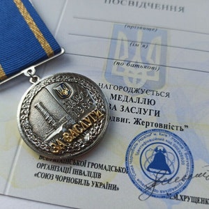 Puede incluir: Medalla plateada con cinta azul y dorada. La medalla presenta un diseño detallado con texto e imágenes ucranianas, incluyendo un escudo y una estructura. El documento adjunto tiene texto y un sello.