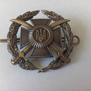 Może przedstawiać: Brązowa odznaka wojskowa z centralnym motywem krzyża z skrzyżowanymi mieczami, otoczona wieńcem laurowym. Odznaka zawiera okrągły emblemat z symbolem trójzębu. Całość jest szczegółowa i bogato zdobiona.