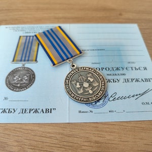 May include: A silver medal with a blue and yellow ribbon, resting on a light blue document. The medal features text in Ukrainian and a design with crossed axes and a hammer. The document also has Ukrainian text.
