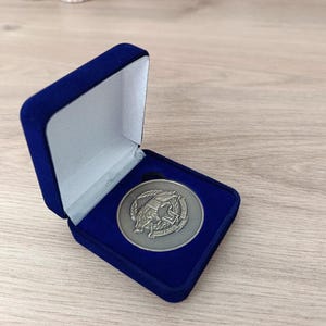 Może przedstawiać: Srebrny medal w otwartym, niebieskim aksamitnym pudełku prezentacyjnym. Medal ma szczegółowy emblemat. Pudełko stoi na drewnianej powierzchni. Projekt medalu jest widoczny. Pudełko jest w kolorze granatowym.
