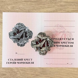 Könnte beinhalten: Zwei Metallabzeichen mit einem zentralen Design, das einen Vogel, ein Gebäude und die Zahl "986" auf einer rosa Karte zeigt. Die Abzeichen sind von einem Kranzdesign umgeben. Text auf Ukrainisch ist auf der Karte zu sehen.