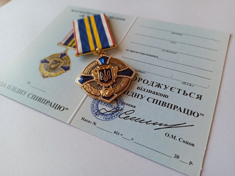 May include: A gold-colored medal with a blue and black cross design, featuring a crest, rests on a light blue document. The medal is accompanied by a ribbon with blue, yellow, and red stripes. The document has Ukrainian text.