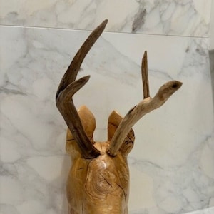Op de afbeelding: Een houten sculptuur van een hertenkop met geweien. De sculptuur is gesneden uit lichtbruin hout en heeft een natuurlijke afwerking.
