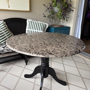 May include: A round pedestal table featuring a black wooden base with three curved legs and a circular tabletop surface covered in a tan and dark brown damask pattern. The table stands on a central column with a smooth, matte black finish.
