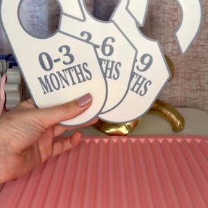 Puede incluir: Separadores blancos de tallas de ropa de bebé con ribete gris, etiquetados 0-3, 3-6 y 6-9 meses. Los separadores se sostienen frente a una bandeja rosa y un objeto dorado.