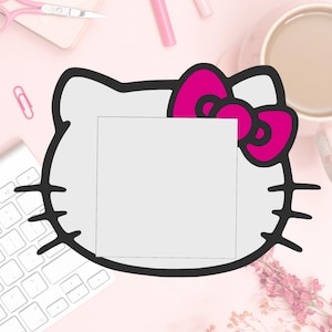 May include: A white Hello Kitty-shaped design with a pink bow and a blank white square in the center. The design is on a pink surface with a keyboard, scissors, and a cup of coffee.