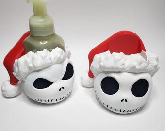 Puede incluir: Dos dispensadores de jabón y soporte blancos con el tema de Jack Skellington. Cada uno tiene un gorro de Papá Noel rojo con ribete blanco. Uno contiene una botella transparente con líquido verde. Las caras tienen ojos negros y bocas cosidas.