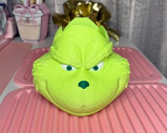 Puede incluir: Un recipiente en forma de Grinch verde lima con una expresión de mal humor, con ojos verdes y una nariz verde oscuro. El recipiente está sobre una superficie rosa, lo que sugiere un artículo festivo o temático.