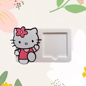 May include: A white rectangular frame with a Hello Kitty character attached. The character is gray with a pink dress and flower. The background has floral and leaf designs.