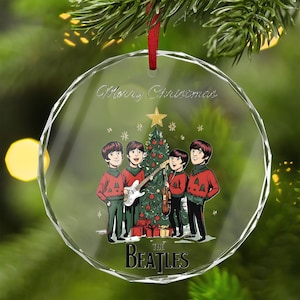 Puede incluir: Un adorno navideño redondo y transparente con una cinta roja. El adorno presenta una imagen de dibujos animados de The Beatles frente a un árbol de Navidad, con las palabras "Merry Christmas" y "The Beatles".