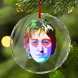 May include: A clear, round Christmas ornament featuring a colorful portrait of a man with round glasses. The image has a rainbow color scheme and the word "Imagine" is written in cursive at the bottom. A red ribbon is attached for hanging.