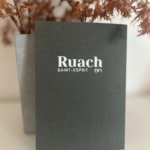 Peut inclure: Une carte gris foncé avec le mot "Ruach" en blanc, ainsi que "SAINT-ESPRIT" et d'autres textes. La carte est présentée devant un arrière-plan flou avec des plantes séchées dans un pot gris.