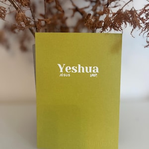 Peut inclure: Un livre rectangulaire chartreuse avec le mot "Yeshua" en blanc, ainsi que les mots "JESUS" et du texte dans une autre langue. Le livre est posé sur un fond flou de branches sèches.