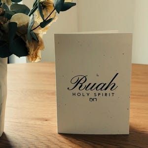 Peut inclure: Une carte crème avec le mot "Ruah" en écriture cursive, avec "HOLY SPIRIT" et du texte hébreu en dessous. La carte est posée sur une table en bois, avec un vase de fleurs séchées en arrière-plan.