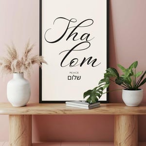 Peut inclure: Une affiche encadrée avec les mots "Tha Com" en écriture élégante, avec "PEACE" et le mot hébreu "Shalom" en dessous. L'affiche encadrée noire est exposée sur une console en bois avec des éléments décoratifs.