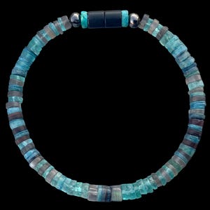 Op de afbeelding: Een cirkelvormige kralenketting met kleine, platte, schijfvormige kralen in verschillende tinten blauw, groenblauw en grijs. De ketting bevat twee metallic zilverkleurige bolvormige kralen en een zwarte cilindrische magnetische sluiting.