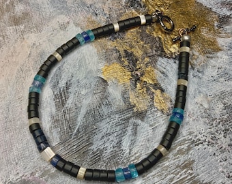 May include: Black beaded bracelet with silver accents and blue and clear glass beads. The bracelet features a toggle clasp and is displayed on a textured background. The bracelet is approximately 7.5 inches in length.