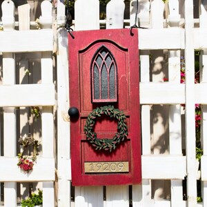 Könnte beinhalten: Rotes, holzförmiges Türschild mit schwarzem Türknauf, grünem Kranz und Fenster mit schwarzem Innenraum. Die Nummer 19209 ist in goldfarbenen Buchstaben geschrieben. Es hängt an einem weißen Zaun.