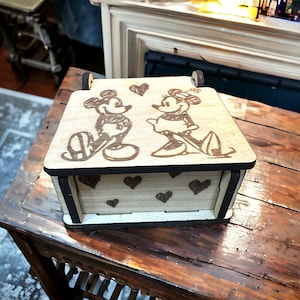 Puede incluir: Una caja de madera con tapa abatible. La tapa presenta un diseño grabado con láser de Mickey Mouse y Minnie Mouse con corazones. La caja está decorada con un patrón de pequeños corazones.