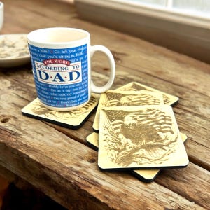 May include: Set of four wooden coasters featuring an American bald eagle with an American flag in the background. The coasters are light brown with a dark brown border. A white mug with blue and red text that reads "The Word According to Dad" is sitting on top of the coasters.