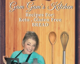 May include: A woman smiles while holding a black sign that says "Eat" in front of a wooden cutting board. The cutting board has the text "Gam Gam's Kitchen Recipes For: Keto. Gluten Free BREAD BY: Nadine J. Larder aka "Gam Gam" written on it.