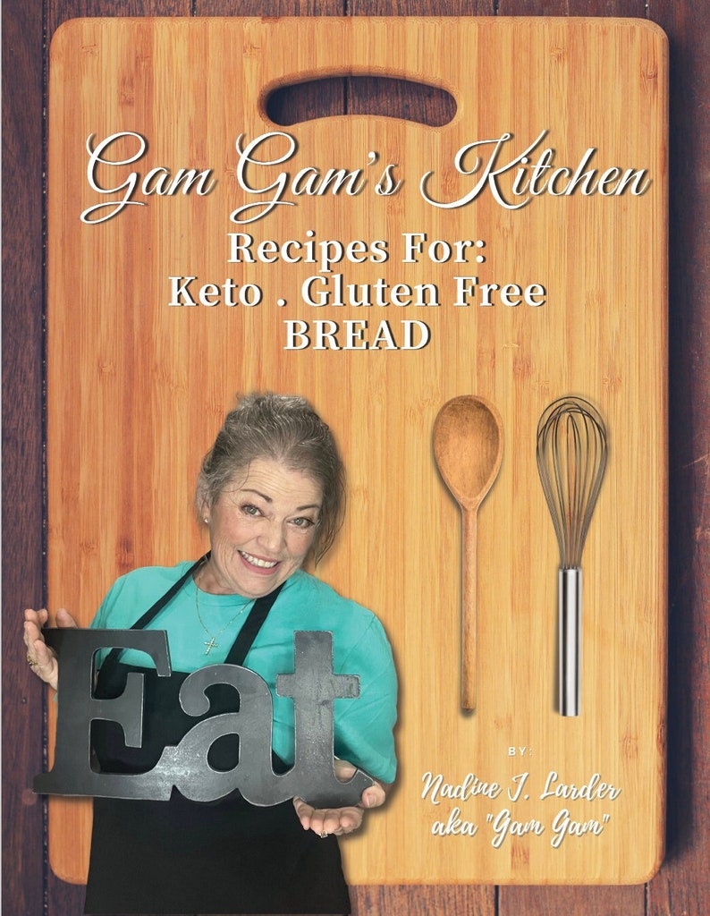 May include: A woman smiles while holding a black sign that says "Eat" in front of a wooden cutting board. The cutting board has the text "Gam Gam's Kitchen Recipes For: Keto. Gluten Free BREAD BY: Nadine J. Larder aka "Gam Gam" written on it.