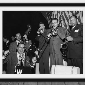 May include: Black and white framed photograph of a jazz band performing on stage. The band includes musicians playing a saxophone, trombone, trumpet, and double bass. The band is in front of an American flag.
