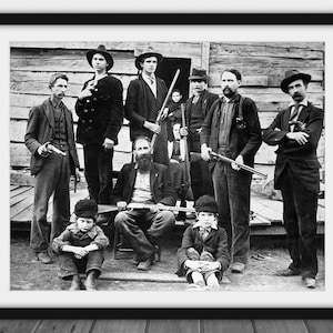 Pode incluir: Impressão emoldurada em preto e branco de um grupo de homens e meninos, alguns segurando rifles e pistolas, posando em frente a uma estrutura de madeira. Fotografia vintage.