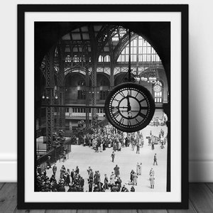 May include: Black and white framed print of a bustling train station interior. A large clock hangs in the foreground, with people milling about below. The architecture features arched ceilings and intricate metalwork.