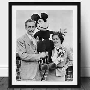 May include: Black and white framed photograph featuring Walt Disney, a woman, and a Mickey Mouse plush toy. The trio is posed outdoors, with the plush toy held between them. The photograph is in a black frame.