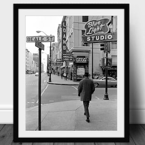 Pode incluir: Fotografia emoldurada a preto e branco de uma cena de rua da cidade. Um homem caminha na calçada em frente a lojas e sinais de rua. Os sinais dizem "LOANS", "BEALE ST." e "Blue Light STUDIO". Carro clássico estacionado.