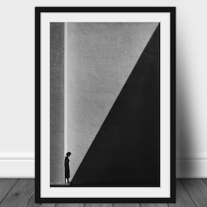 Può includere: Stampa artistica incorniciata in bianco e nero con una composizione architettonica minimalista. Una figura silhouette si trova in uno spazio stretto e illuminato tra due muri, con una grande ombra triangolare. L'opera è incorniciata in nero.