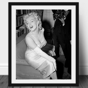 May include: Black and white framed print featuring a smiling woman in a light-colored dress and long gloves. She is seated on a sofa, with a bookshelf in the background. A photographer with a vintage camera is visible.