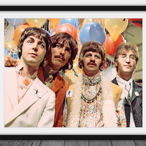 May include: Framed print featuring The Beatles, with the band members posing in front of colorful balloons. The image shows the band members wearing various outfits, including a white suit, a yellow jacket, and a patterned coat. The image is framed in a black border.