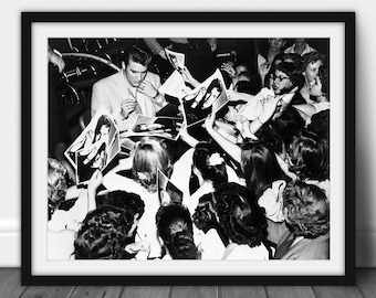 May include: Framed black and white print depicting Elvis Presley signing autographs for a crowd. The photograph shows fans reaching out for signed pictures. A vintage image, ideal for music memorabilia enthusiasts. The print is framed in a black border.
