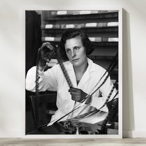 May include: Monochrome framed print showing a woman in a lab coat inspecting film strips. She wears gloves and sits at a table with film reels. The background has shelves with film canisters, giving a vintage feel.