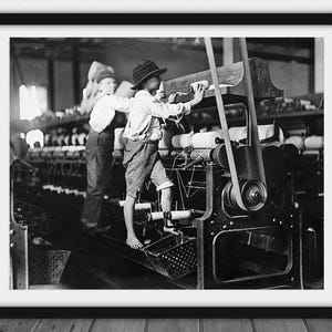 May include: Black and white framed photograph of two young boys working in a textile mill. One boy is standing on a machine, while the other is standing behind him. The photograph captures the industrial era.
