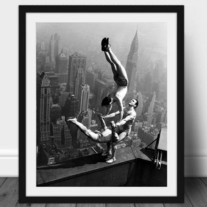 May include: Black and white framed print of three acrobats performing a balancing act on a rooftop with a cityscape in the background. The acrobats are wearing athletic attire.