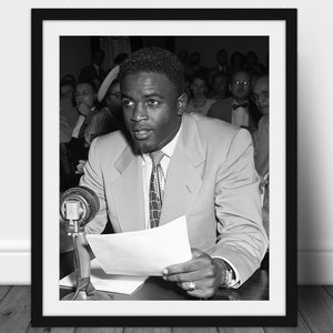 Pode incluir: Impressão emoldurada em preto e branco de um homem de terno falando em um pódio. O homem está segurando um papel e olhando para frente. A impressão está em uma moldura preta e é exibida em uma parede branca. O homem está usando um terno de cor clara e uma gravata estampada.