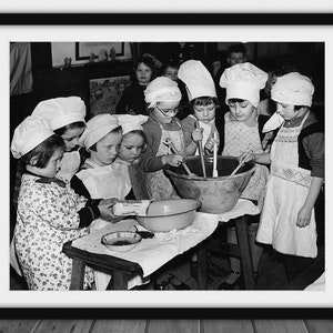 May include: Black and white framed print of children in chef hats and aprons gathered around a large bowl, stirring ingredients. A vintage photograph depicting a cooking class or baking activity.