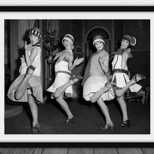 May include: Black and white framed print featuring four women in flapper attire, dancing. They wear short skirts, hats, and heels, with arms raised and legs kicking. The image captures a vintage, theatrical scene, likely from the 1920s.