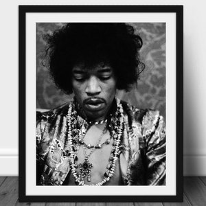 May include: A monochrome framed portrait of a person with a large afro, adorned with a patterned jacket and multiple necklaces. The person is looking down, with a thoughtful expression. The image is a close-up shot.