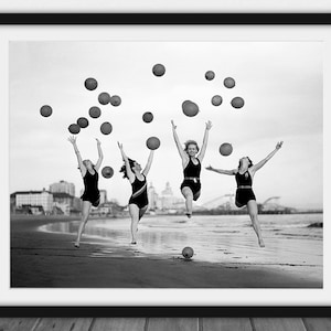 Könnte beinhalten: Schwarz-Weiß-Druck in einem Rahmen, der vier Personen an einem Strand zeigt, die springen und rote Bälle in die Luft werfen. Sie tragen Vintage-Badeanzüge. Der Hintergrund zeigt einen Strand und Gebäude.