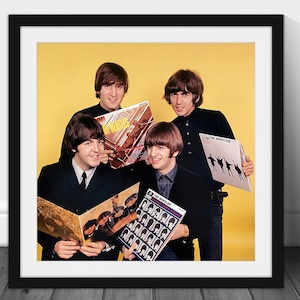 May include: A framed print featuring The Beatles holding their album covers. The band members are dressed in black suits and shirts, with the album covers in the foreground. The background is a solid yellow color.