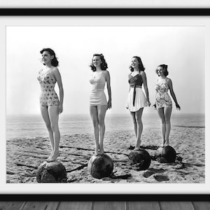 Könnte beinhalten: Schwarz-Weiß-Druck mit Rahmen, der vier Frauen in Vintage-Badeanzügen zeigt, die auf großen, runden Objekten an einem Strand stehen. Die Frauen lächeln, mit dem Meer im Hintergrund.