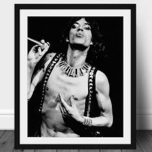 Puede incluir: Impresión enmarcada en blanco y negro de un hombre con cabello oscuro, que lleva un chaleco de cuero con tachuelas y un collar grande. Sostiene un micrófono y tiene una pulsera en la muñeca. La imagen es un retrato.