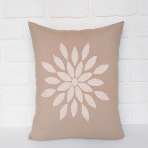 May include: A rectangular cushion in a beige hue, featuring a central floral design in off-white. The flower is composed of numerous petal shapes. The cushion is likely made from a soft material, suitable for home decor.