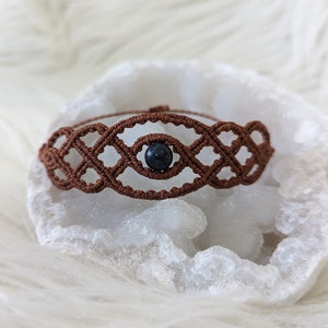 Peut inclure: Un bracelet en macramé marron avec une perle bleu foncé au centre. Le bracelet est sur un fond blanc.