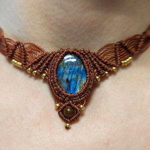 Peut inclure: Un collier ras de cou en macramé marron avec une grosse pierre de labradorite bleue et des perles dorées.